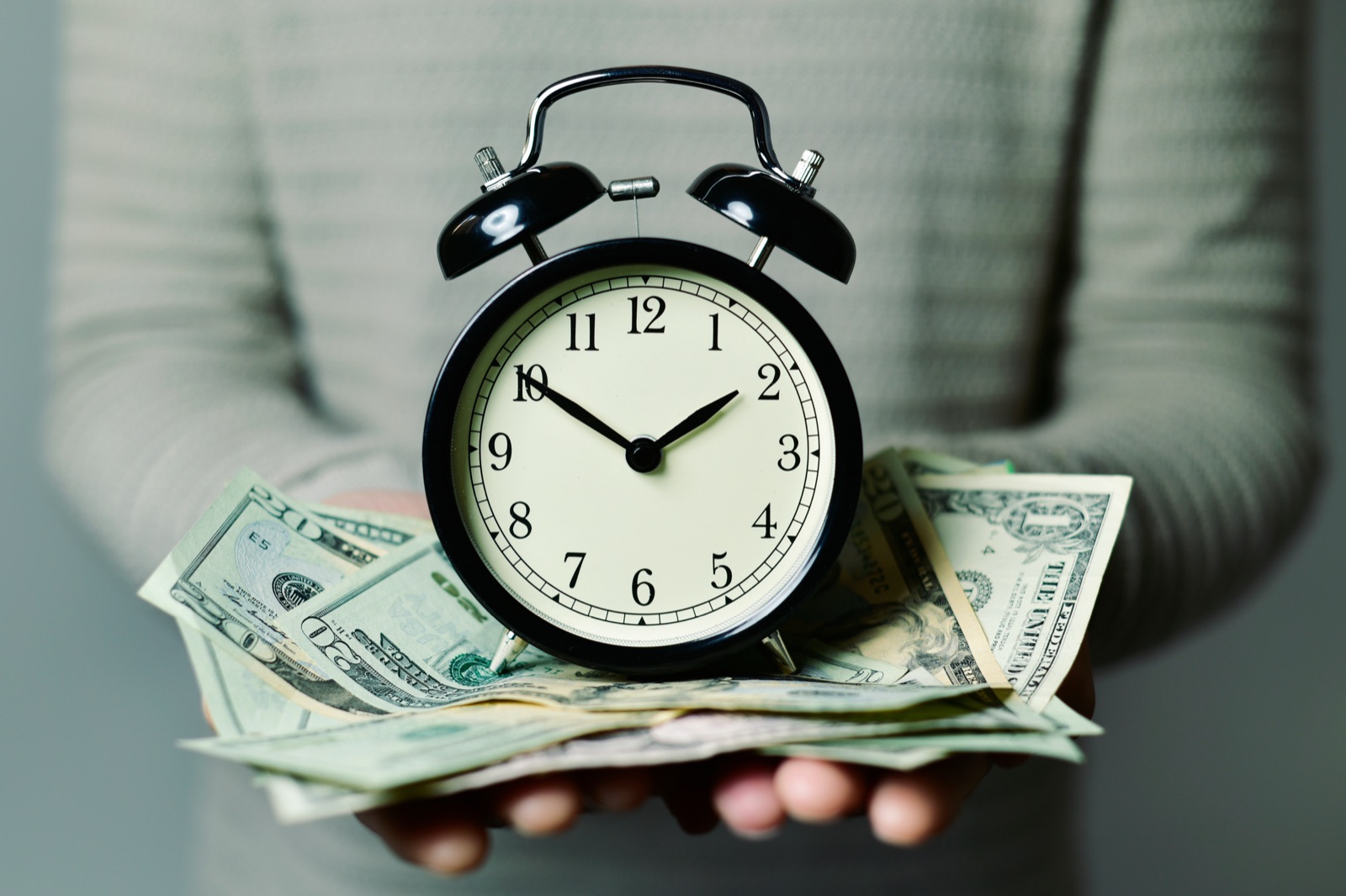 Hands holding an alarm clock resting on a fan of cash